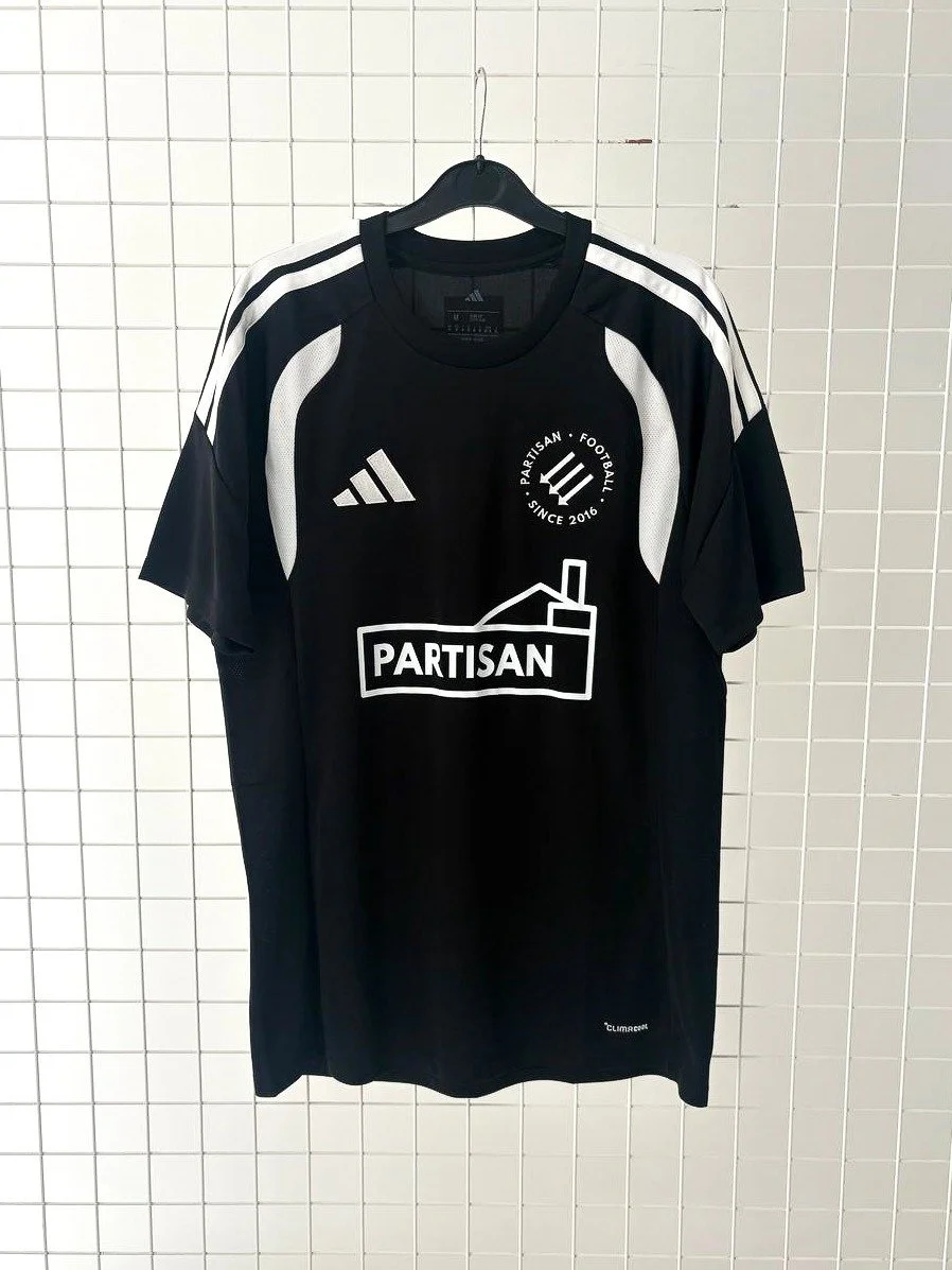 Photo of the front of a Partisan football shirt. The shirt is black with white accents including the Partisan logo in the "sponsor" position and embroidered Adidas and Partisan logos.