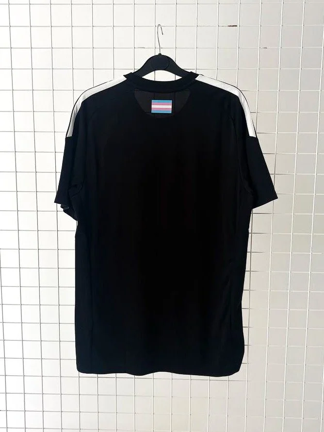 Photo of the back of a Partisan football shirt. The shirt is black with white accents including a trans pride flag on the nape of the neck.