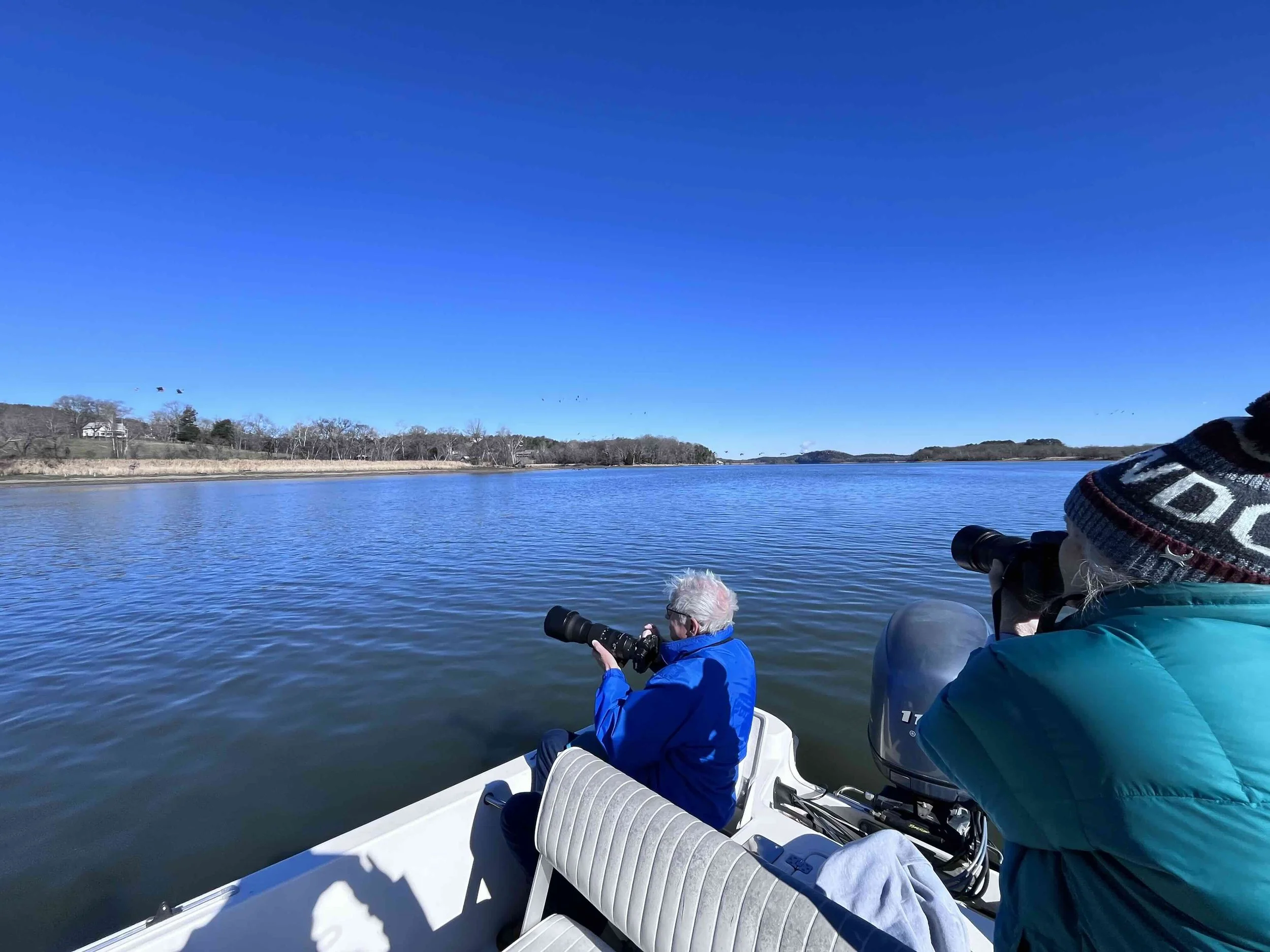 Two people on a boat taking photos of a river with clear blue sky and distant shoreline.