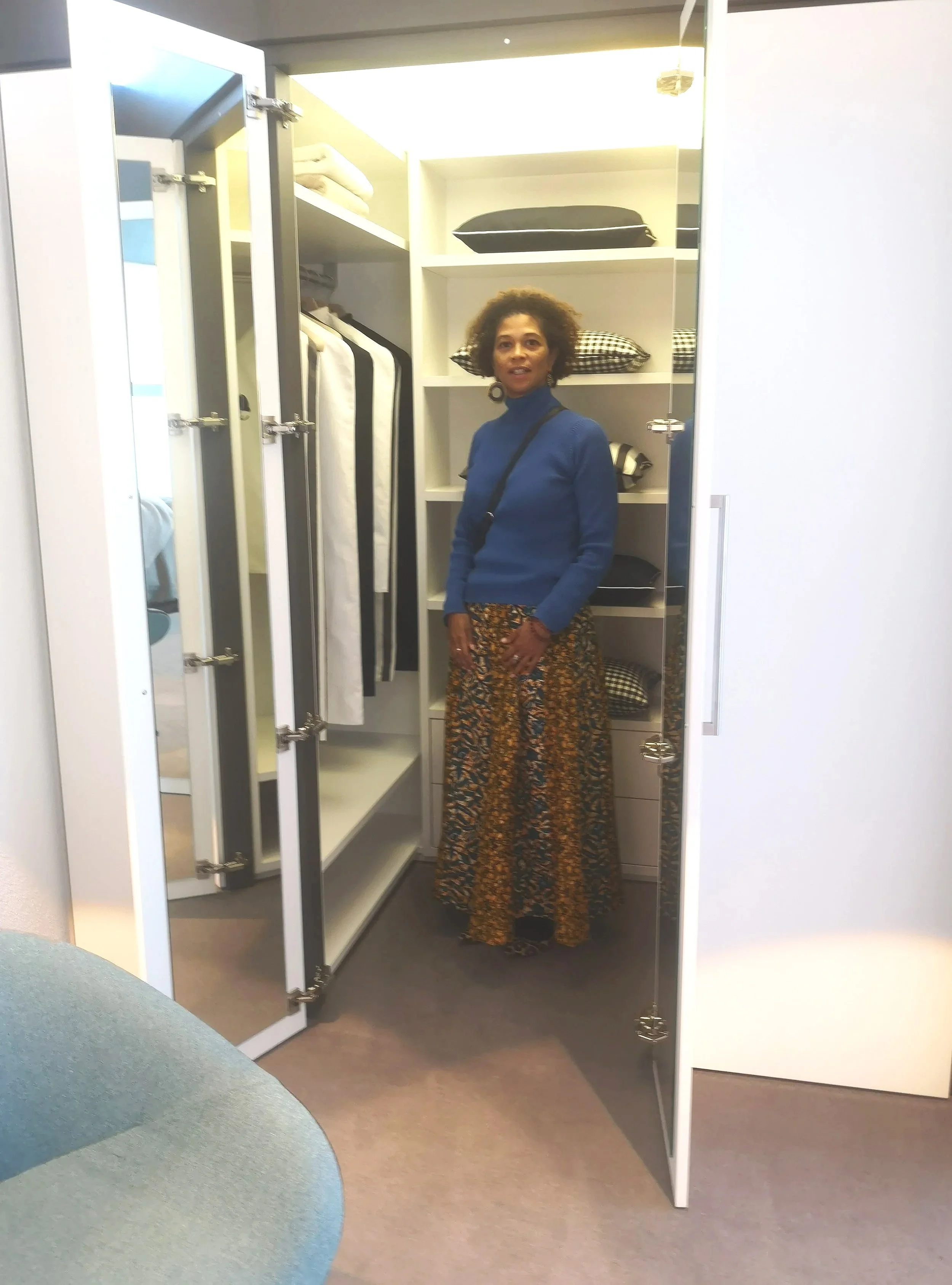 A woman standing inside a walk-in closet with shelves of clothing and pillows, wearing a blue top and a long, patterned skirt.