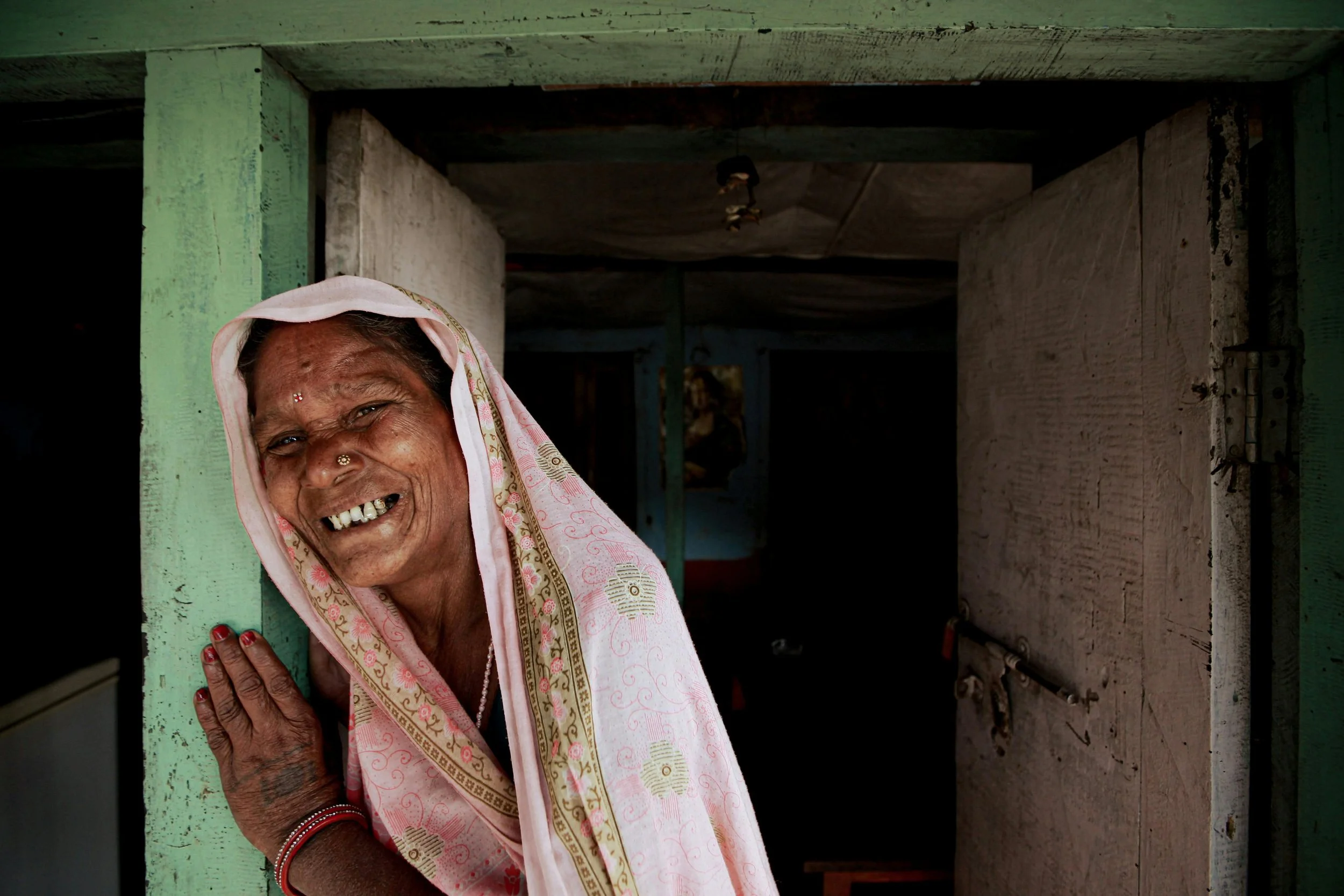 An elderly woman smiling and peeking out from a doorway, wearing a pink saree with gold embroidery, with a joyful expression.