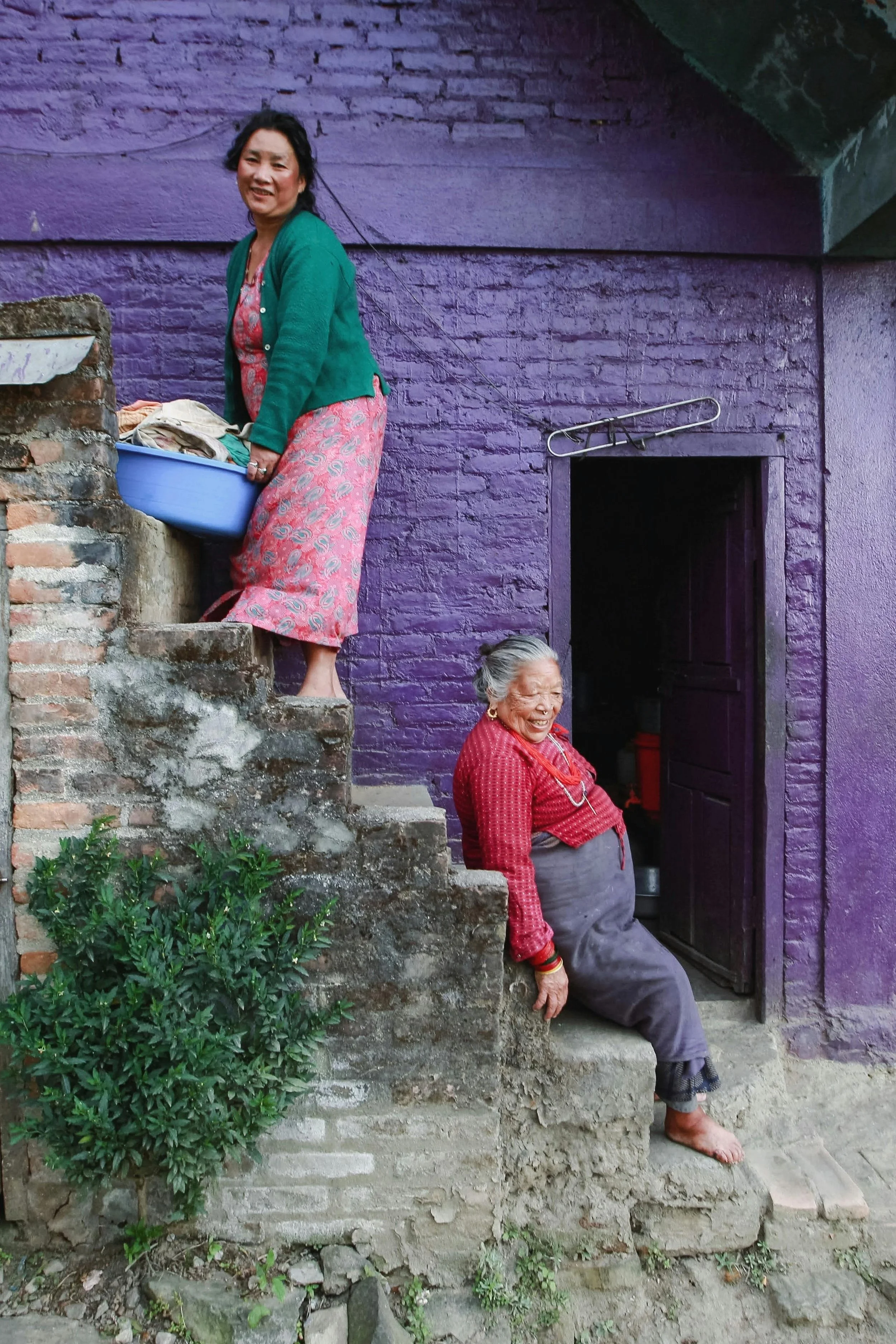 Two women, one middle-aged and one elderly, are on stairs outside a purple-painted brick building. The middle-aged woman is standing on the stairs holding a blue basin with laundry, wearing a pink dress and green cardigan, smiling. The elderly woman is sitting on the bottom step, laughing, wearing a red shirt and gray skirt, with her shoes removed, leaning back slightly.