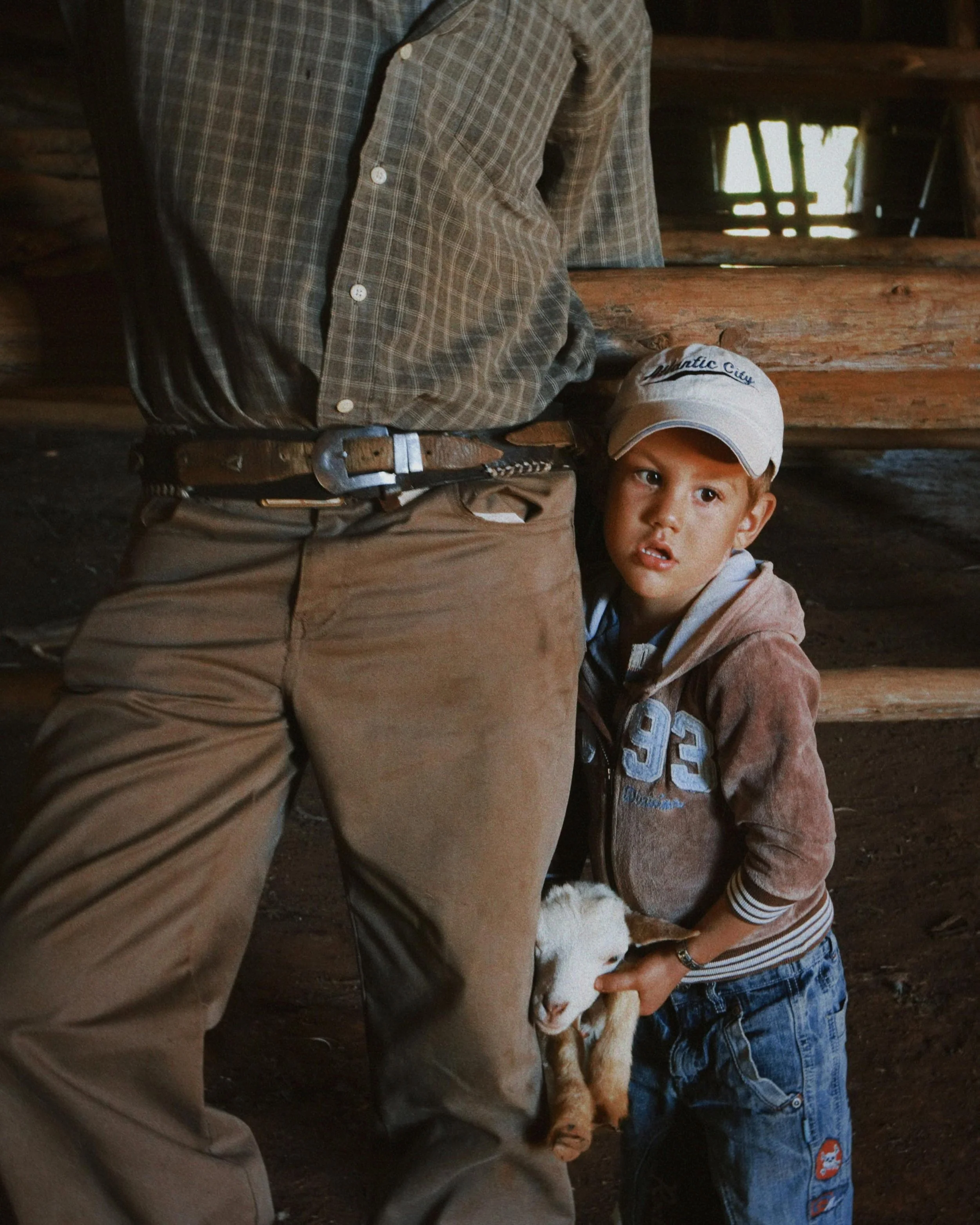 Young boy leaning against an adult's leg, holding a small white and brown goat, inside a rustic wooden barn.