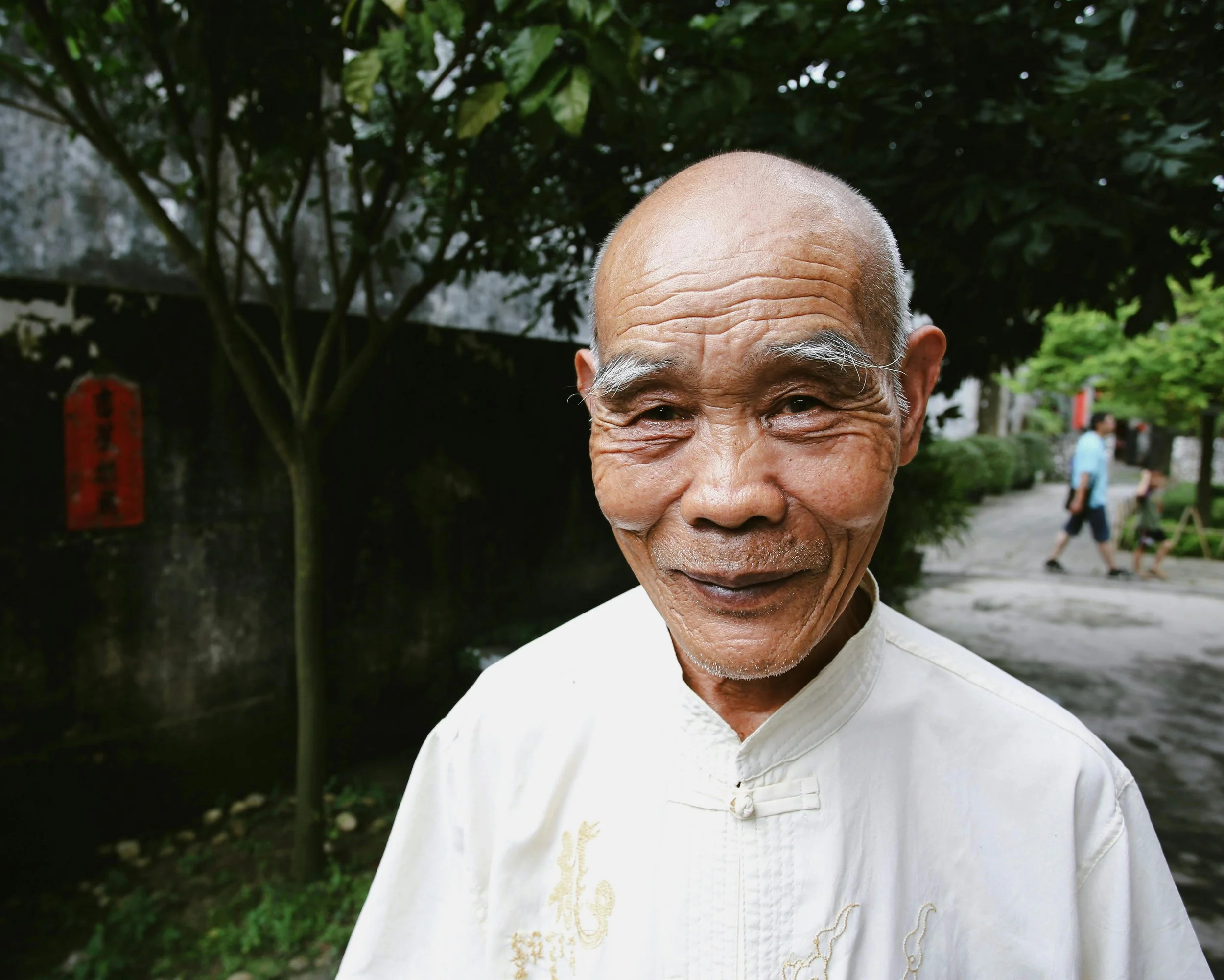 Close-up of an elderly Asian man smiling outdoors, wearing a traditional white shirt with embroidery, with trees and people walking in the background.