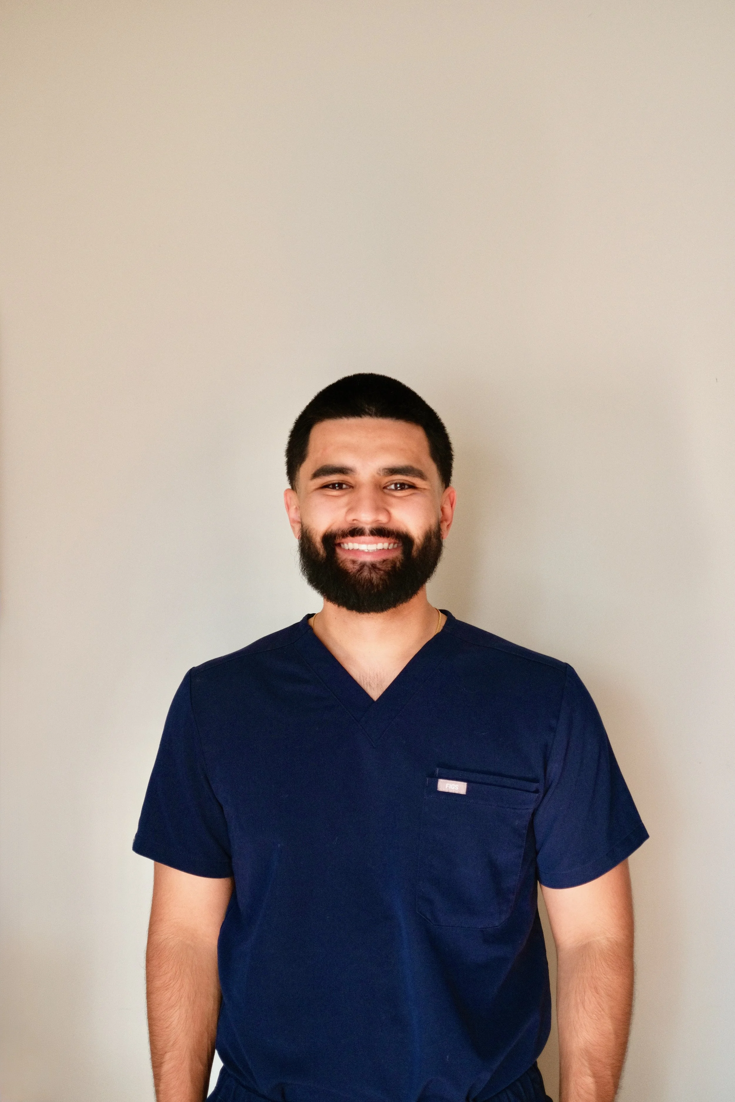 Smiling man in navy scrubs standing against beige wall.
