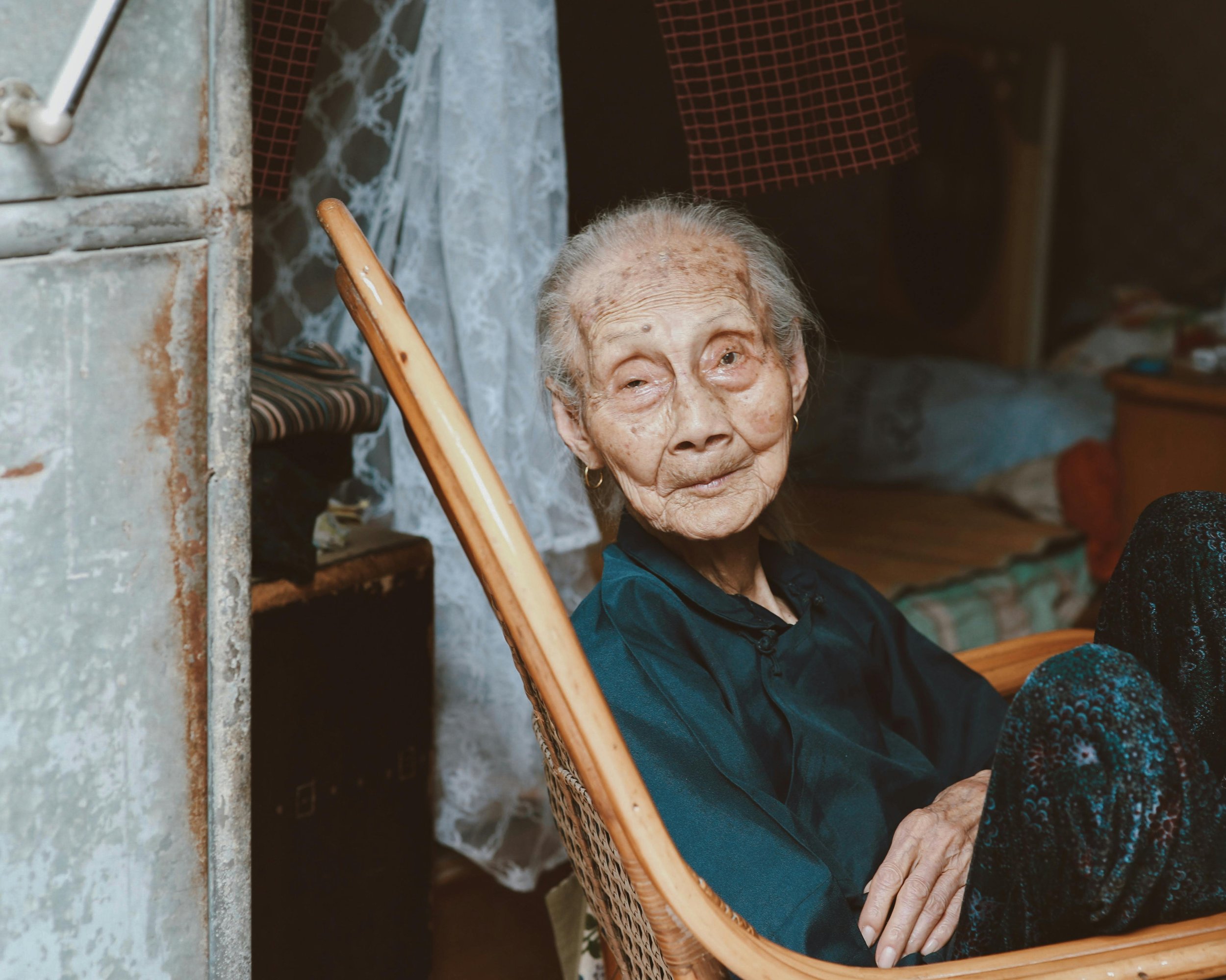 An elderly woman with gray hair and a dark blue shirt sitting in a wicker chair inside a rustic room, looking at the camera with a gentle expression.