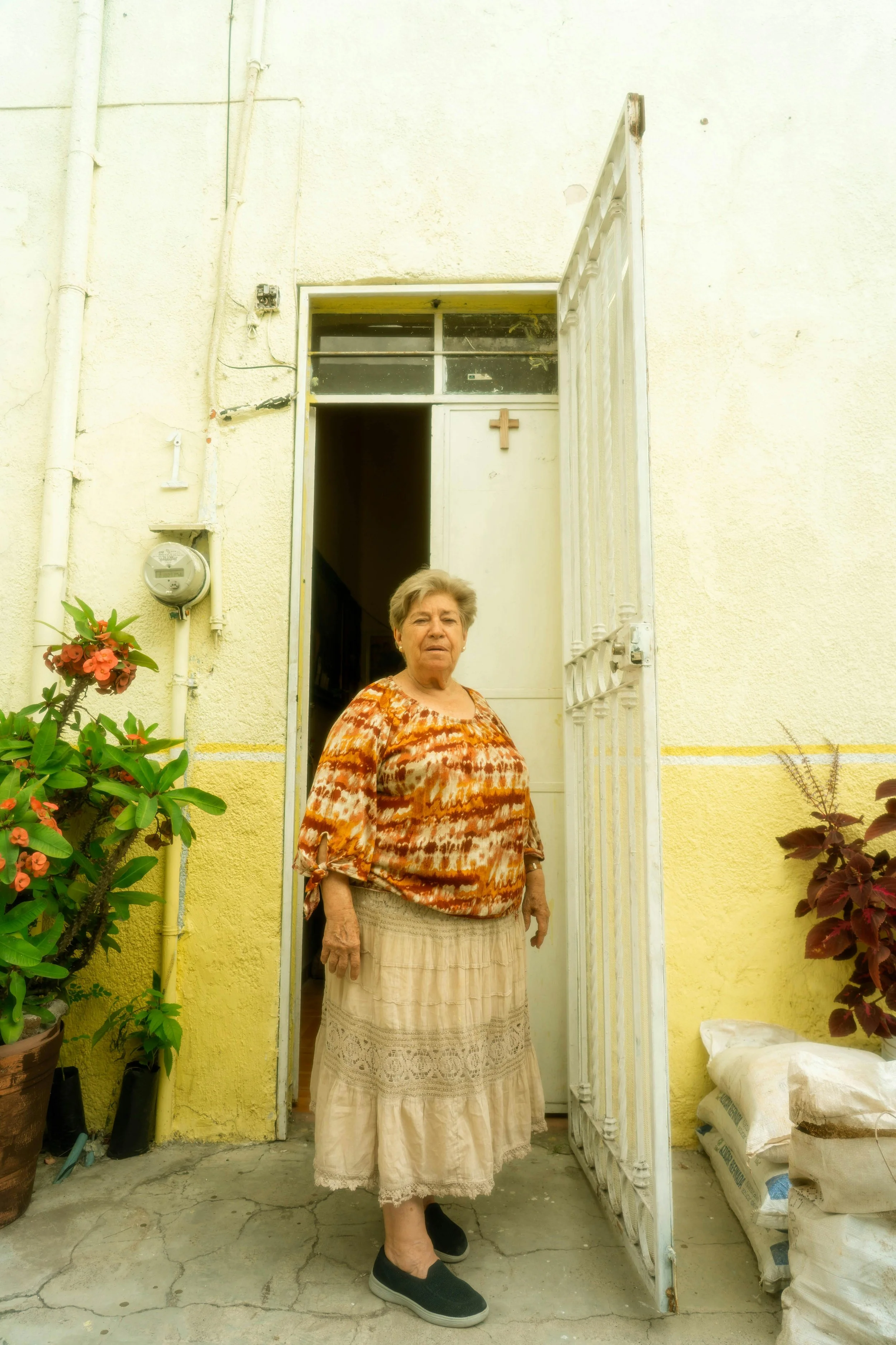 An elderly woman standing in front of a yellow house doorway with a clock, potted plants, and a cross on the door. She wears a patterned top, a long skirt, and slip-on shoes.