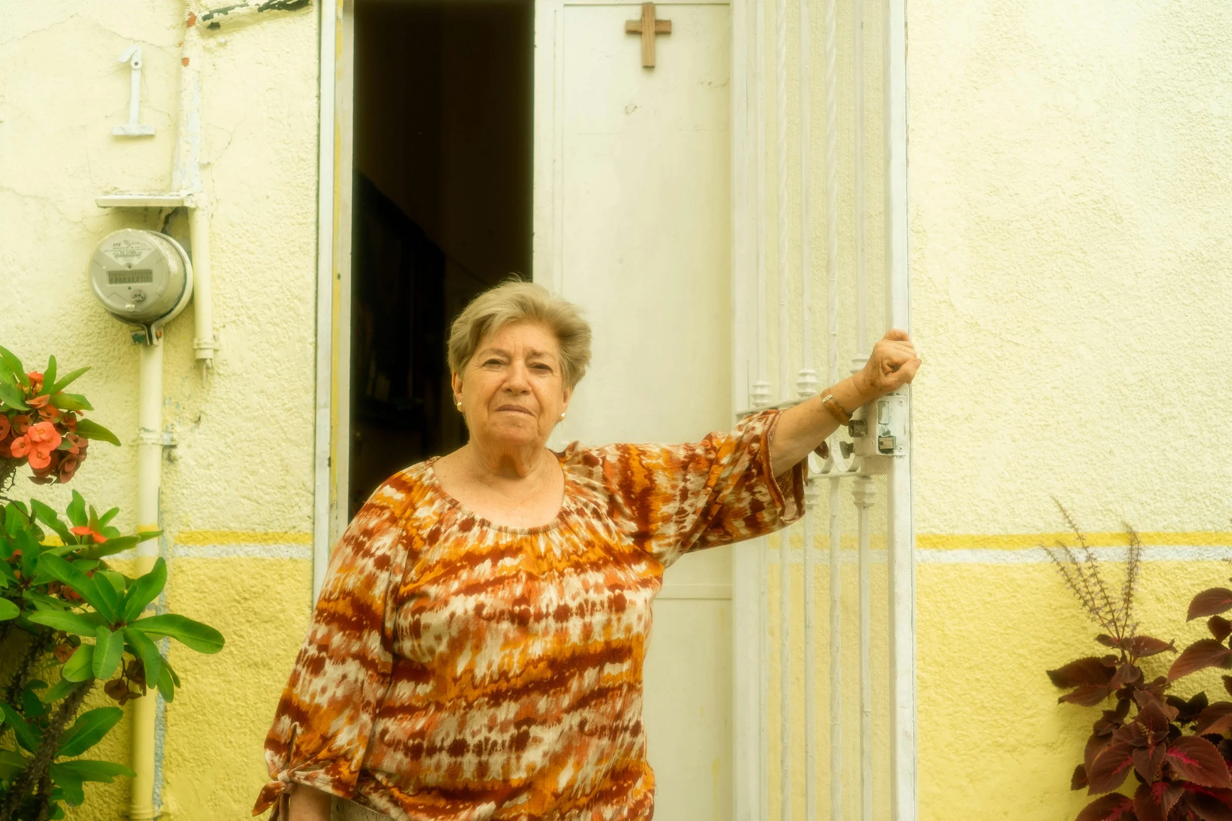 An elderly woman standing outside a yellow house, holding the door open. She is wearing a patterned orange, brown, and white blouse, and has short gray hair.