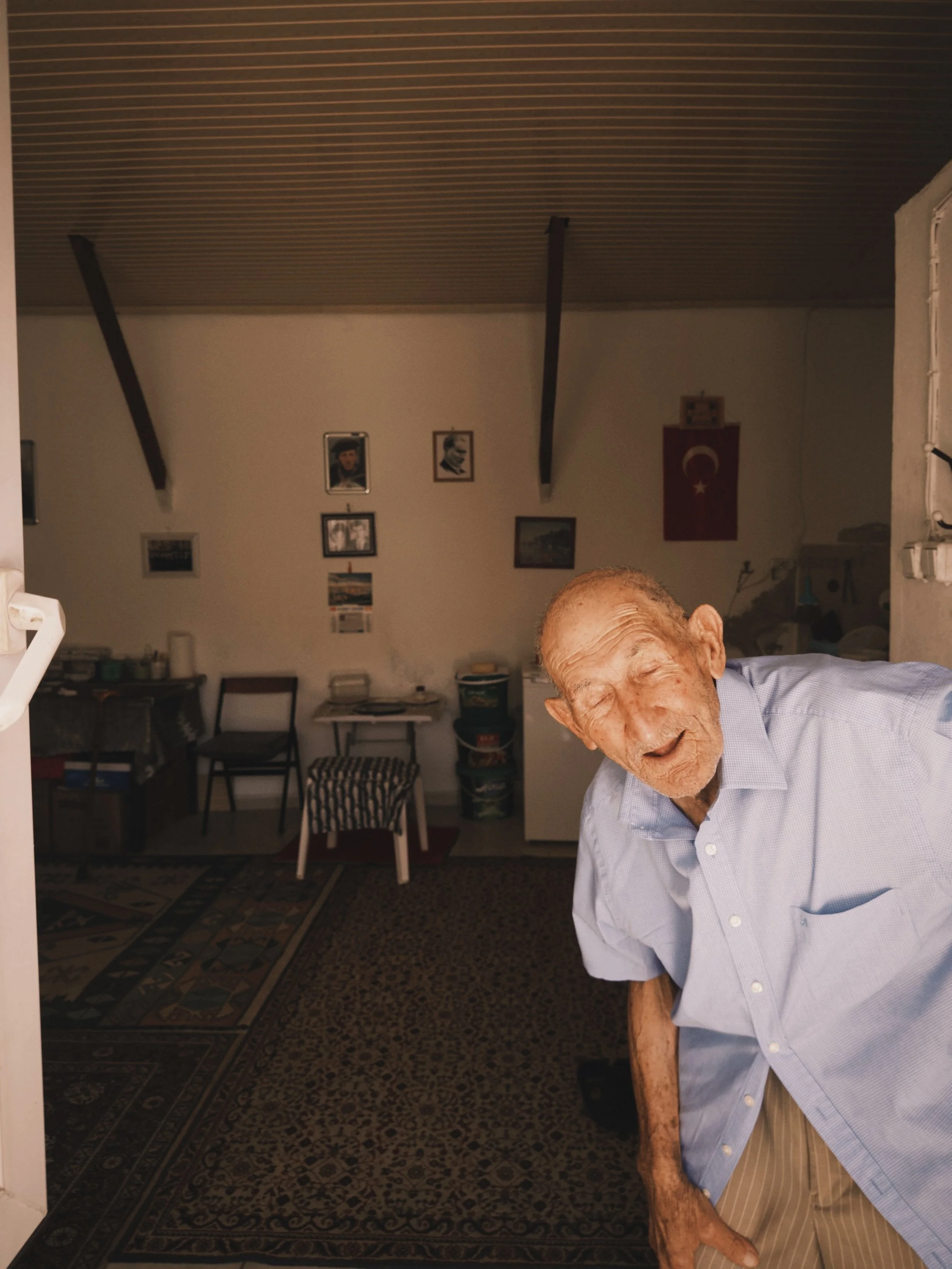 An elderly man with a big smile, wearing a light blue shirt, leaning slightly forward inside a home with Turkish flags and family photos on the wall.