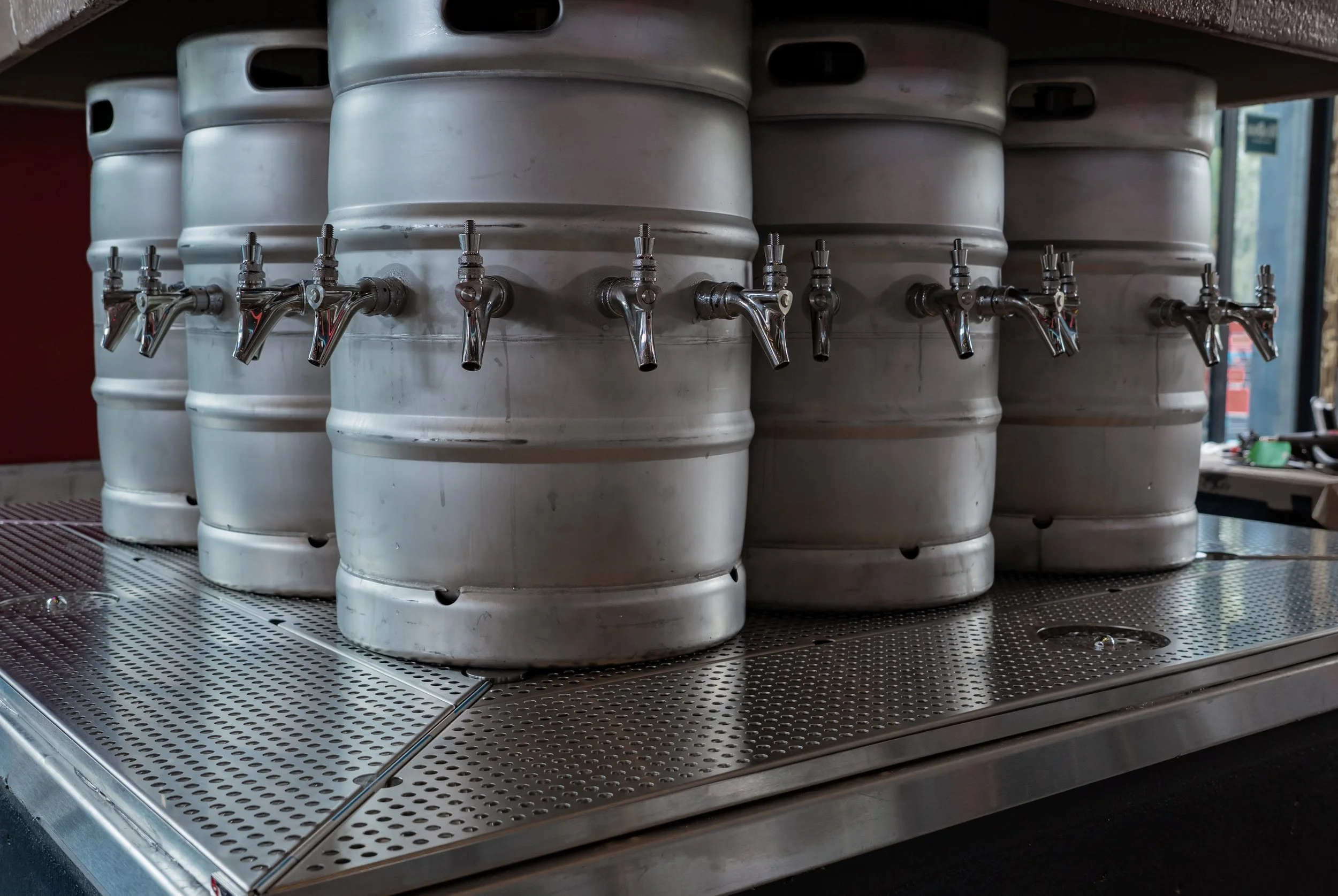 Several white kegs with chrome taps are placed on a metal tray underneath a wooden shelf.