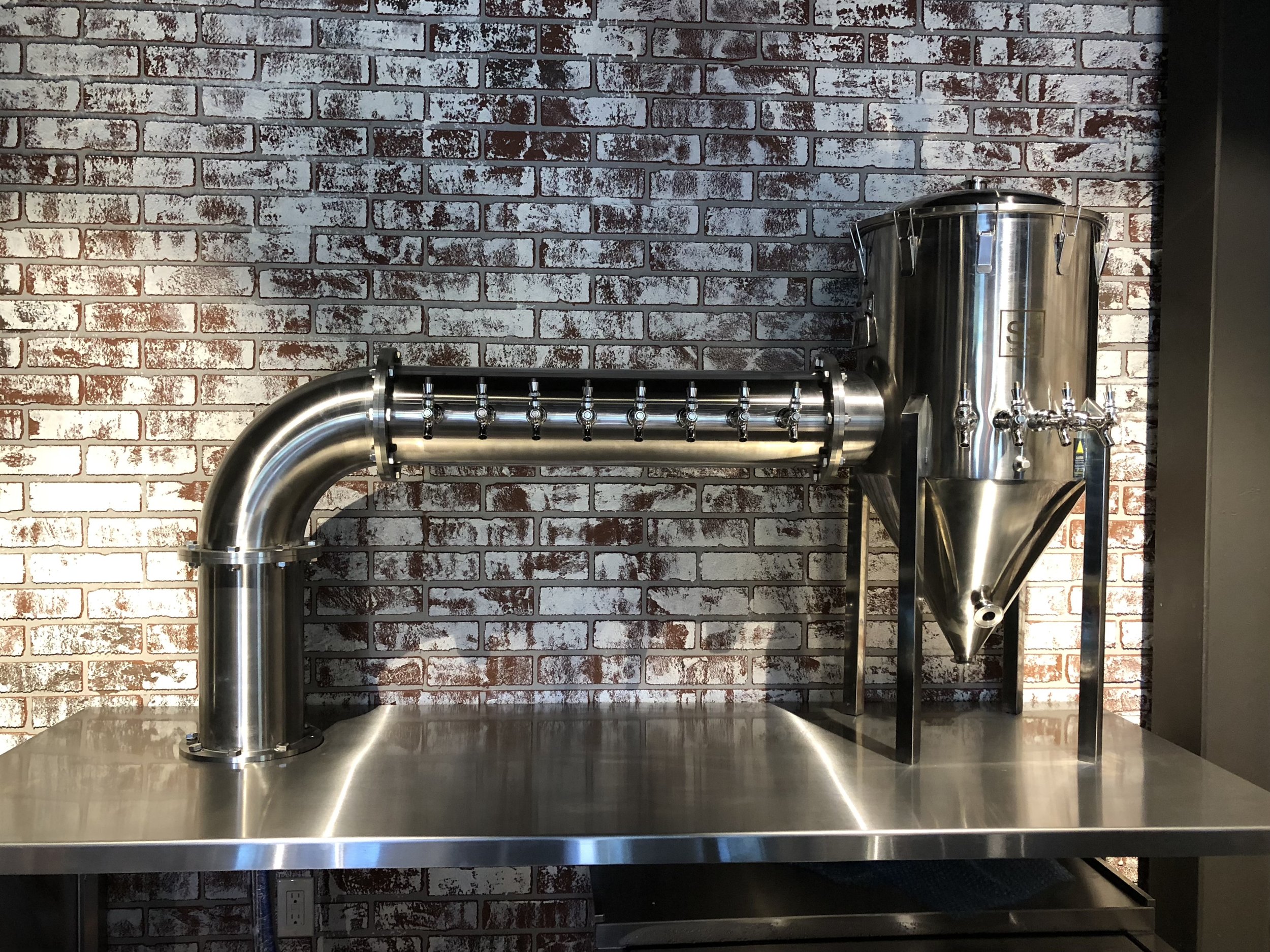 Stainless steel brewing or distillation equipment with pipes and valves against a brick wall background.