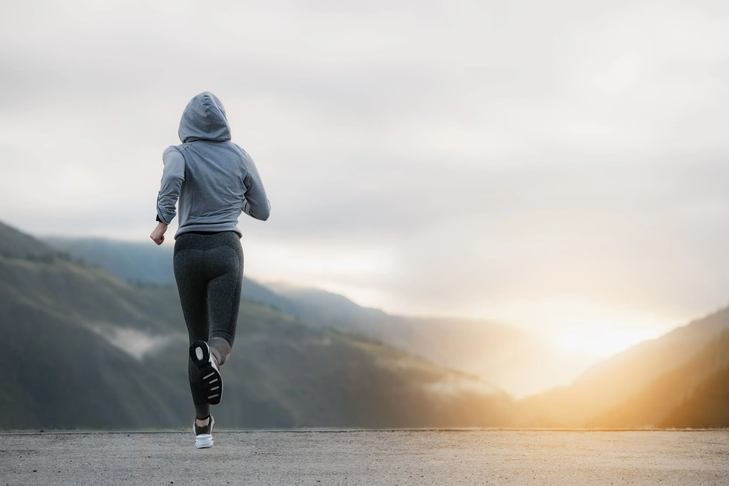 Person jogging on a mountain road at sunrise, wearing a hoodie and leggings.