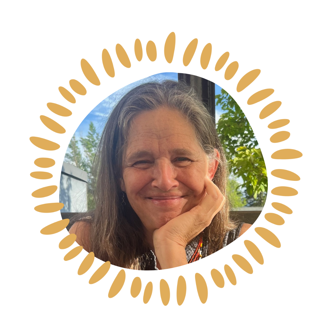 A smiling woman with long brown hair resting her chin on her hand, with a window and greenery behind her.