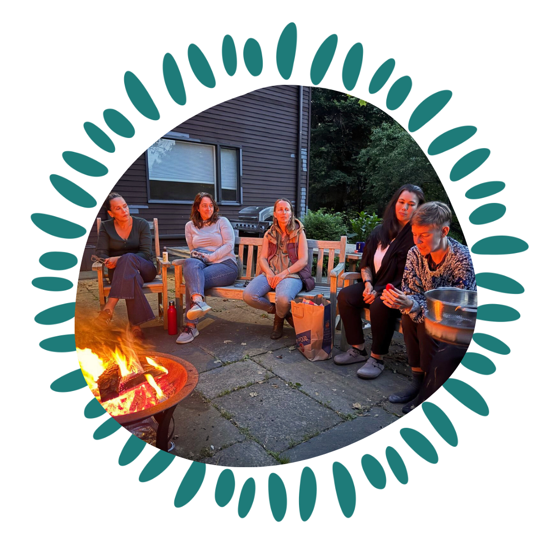 Five women sitting around a fire pit outdoors during evening. One woman is looking at her phone, others are talking or looking around. There are bags and a grill in the background.