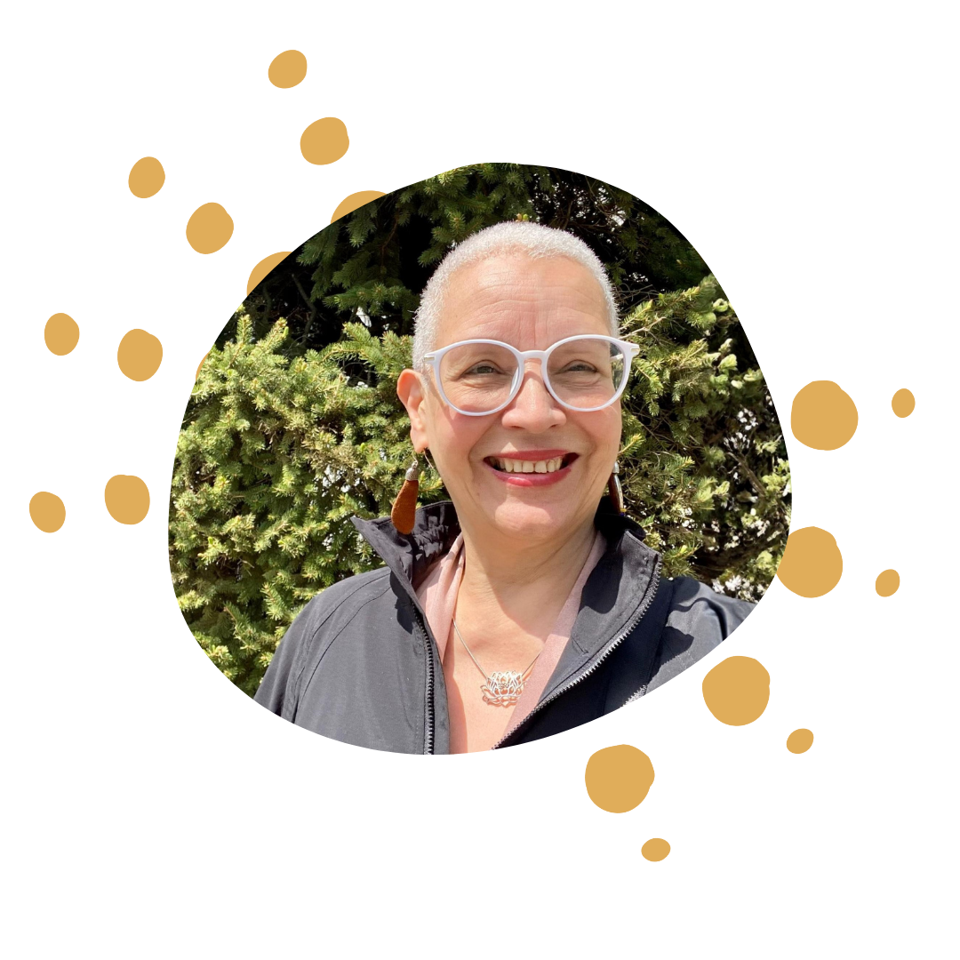 A woman smiling outdoors in front of trees, wearing glasses, earrings, and a jacket.