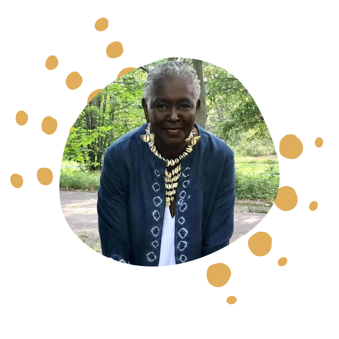 Older woman with gray hair wearing a blue jacket and shell necklace, outdoors in a wooded area.