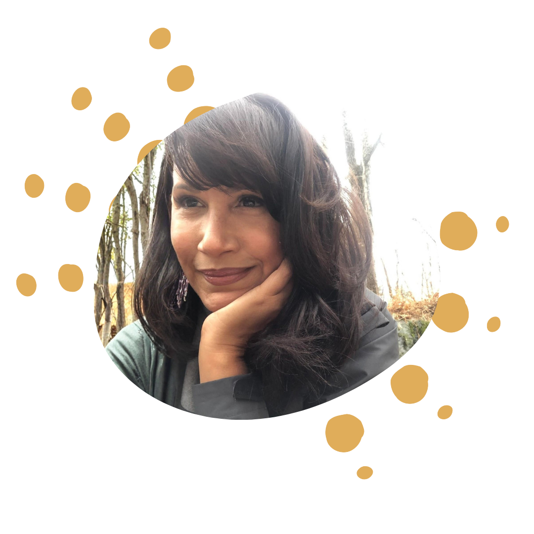 Close-up of a woman with dark hair and makeup, resting her face on her hand outdoors surrounded by trees.