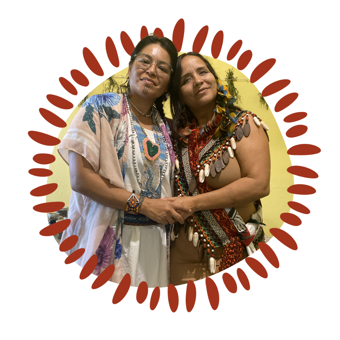 Two women dressed in traditional Indigenous attire, holding hands, standing against a yellow wall, surrounded by decorative red shapes.