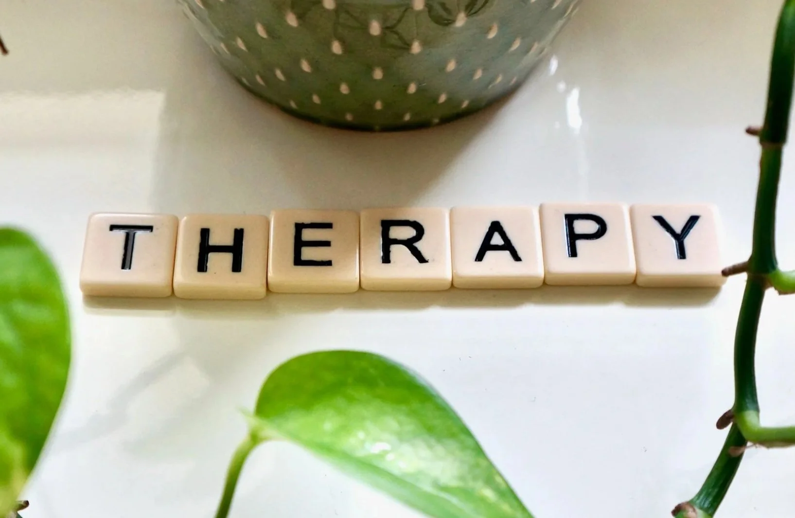 Scrabble tiles spelling out 'THERAPY' arranged on a white surface with green leaves and a decorative bowl.