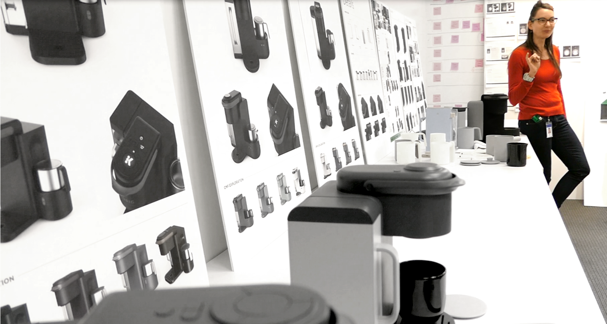 Woman in red sweater standing near a display of various gadgets and accessories on a table and wall in an office or retail setting.