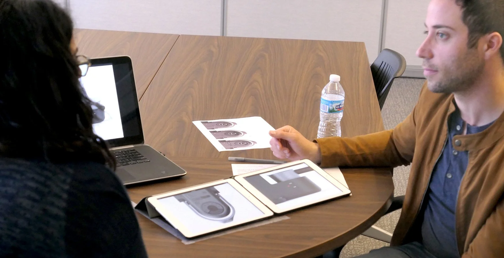 Two people seated at a wooden conference table, engaged in discussion with documents, a laptop, and a tablet, in an office setting.