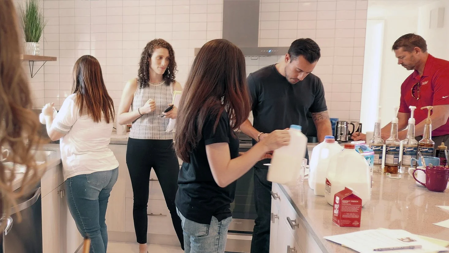 Group of five people in a kitchen, two women and three men, preparing drinks and engaging in tasks.