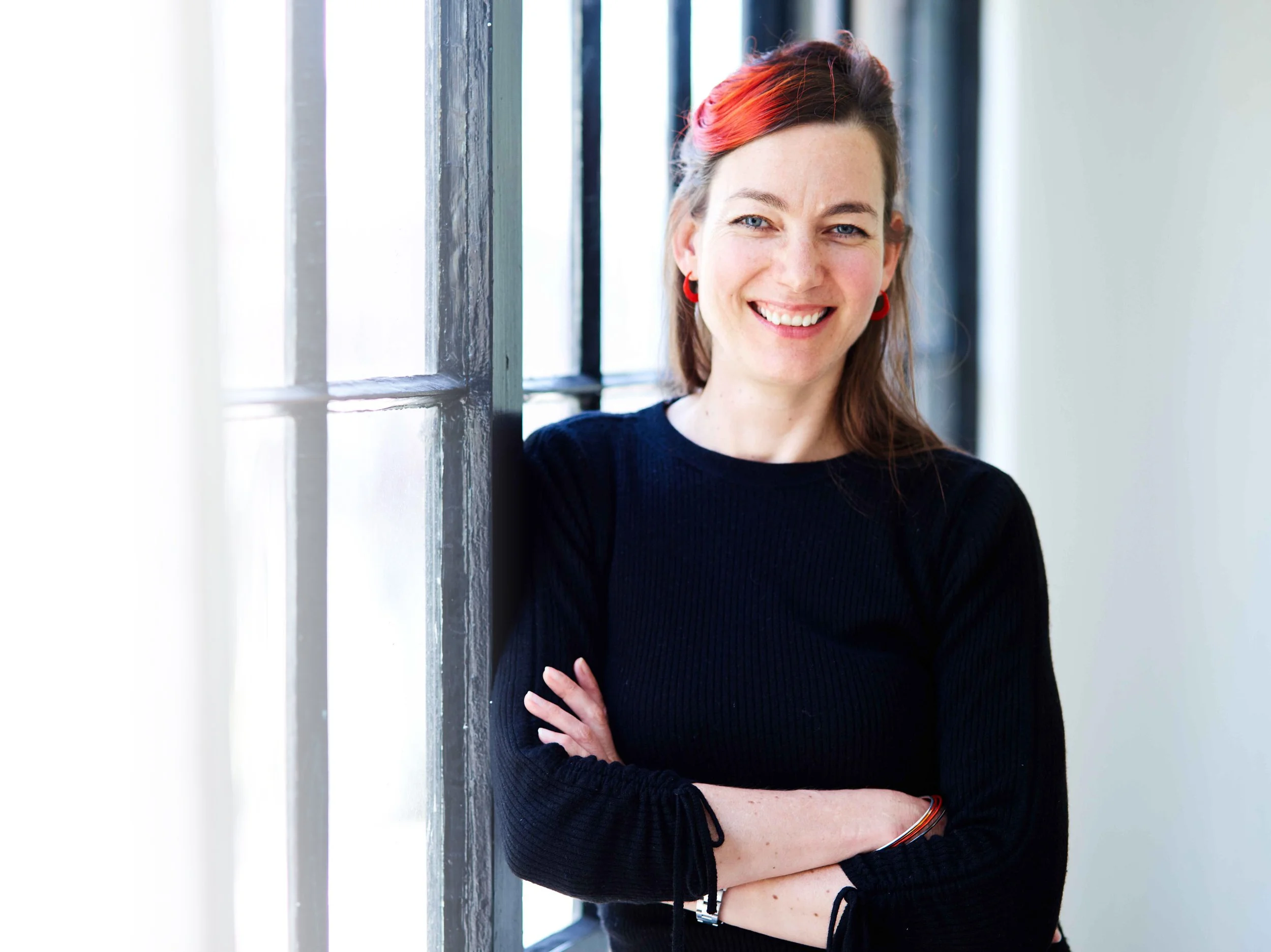 A woman with red and brown hair, wearing a black sweater and red earrings, smiling and standing by a window with crossed arms.