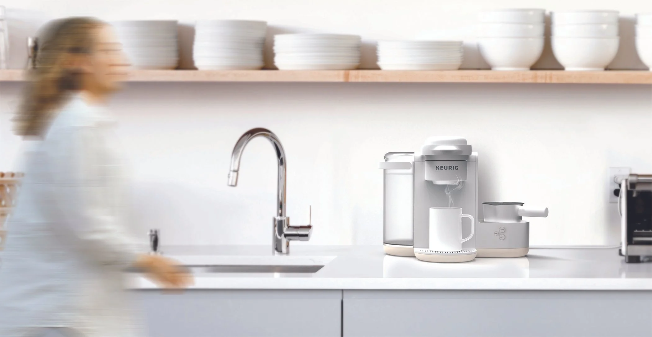 Person in a white outfit walking in a minimalist kitchen with a white countertop, a modern kitchen faucet, a Keurig coffee machine, a white mug, and open shelves with stacked white plates and bowls.