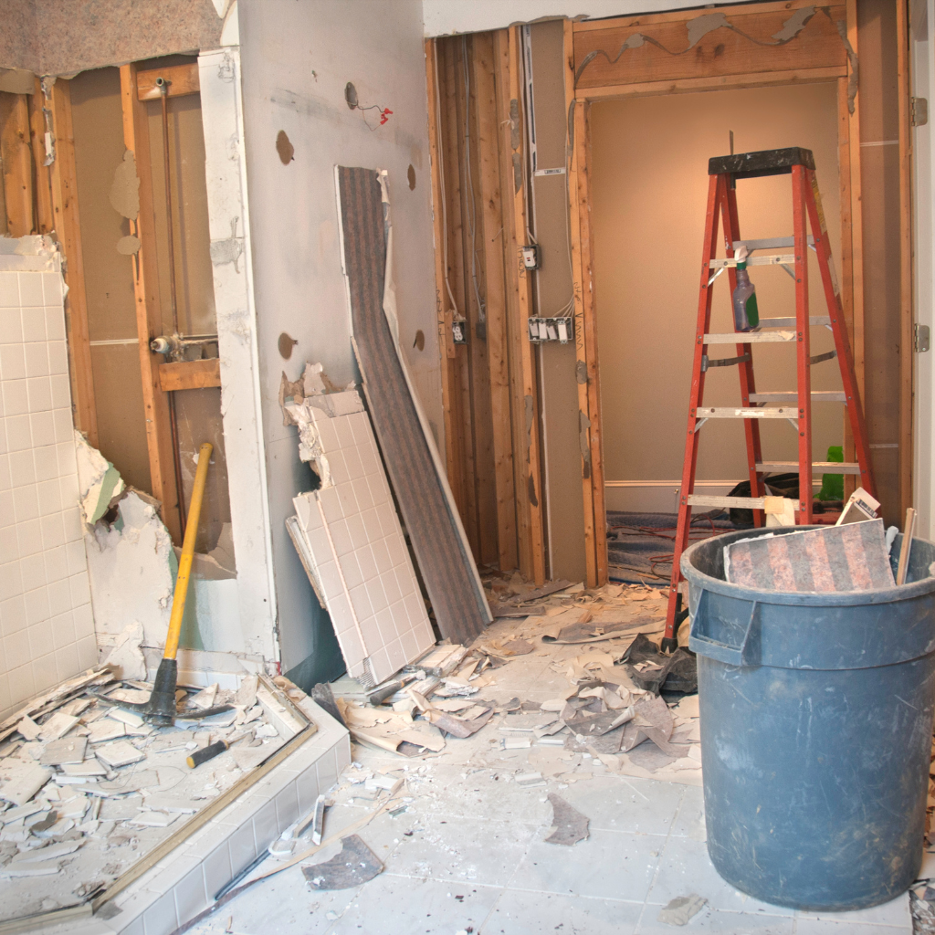 A bathroom under renovation with drywall partially removed, debris on the floor, a ladder, and construction tools.
