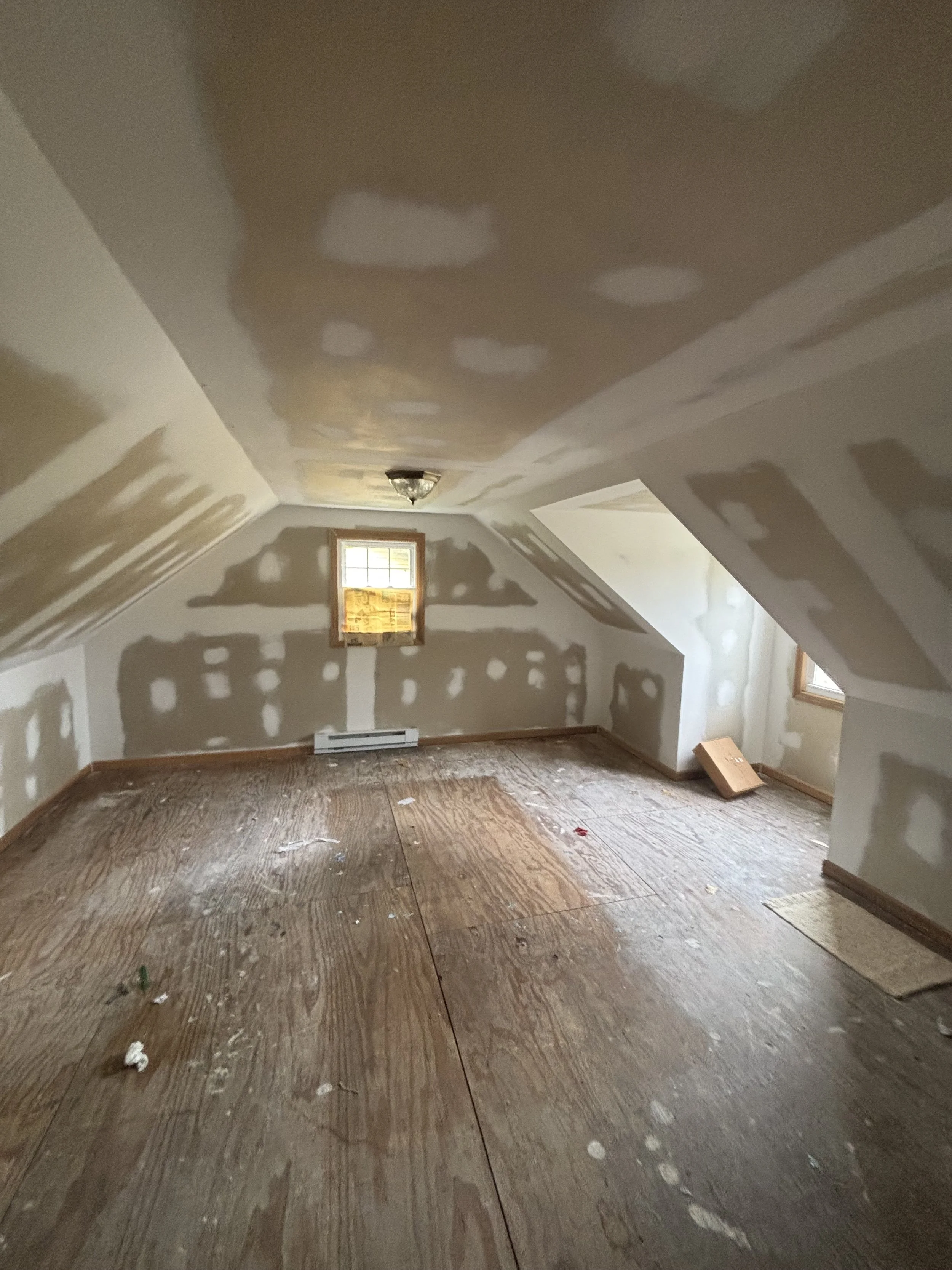 Unfinished attic room with drywall installation and patching, with wooden floors, small windows, and a ceiling light fixture.
