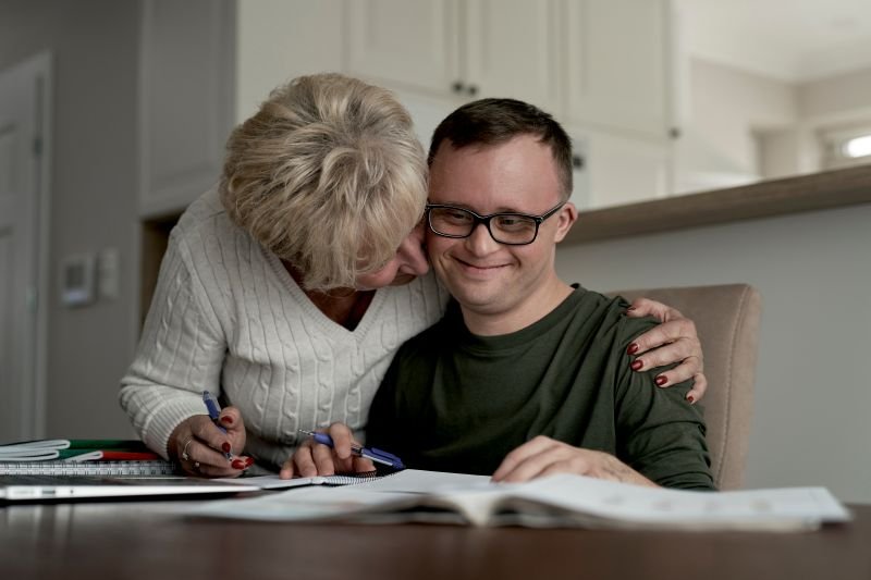 Older woman wearing a white sweater hugging younger man with down syndrome who is seated at a table with papers in front of him.