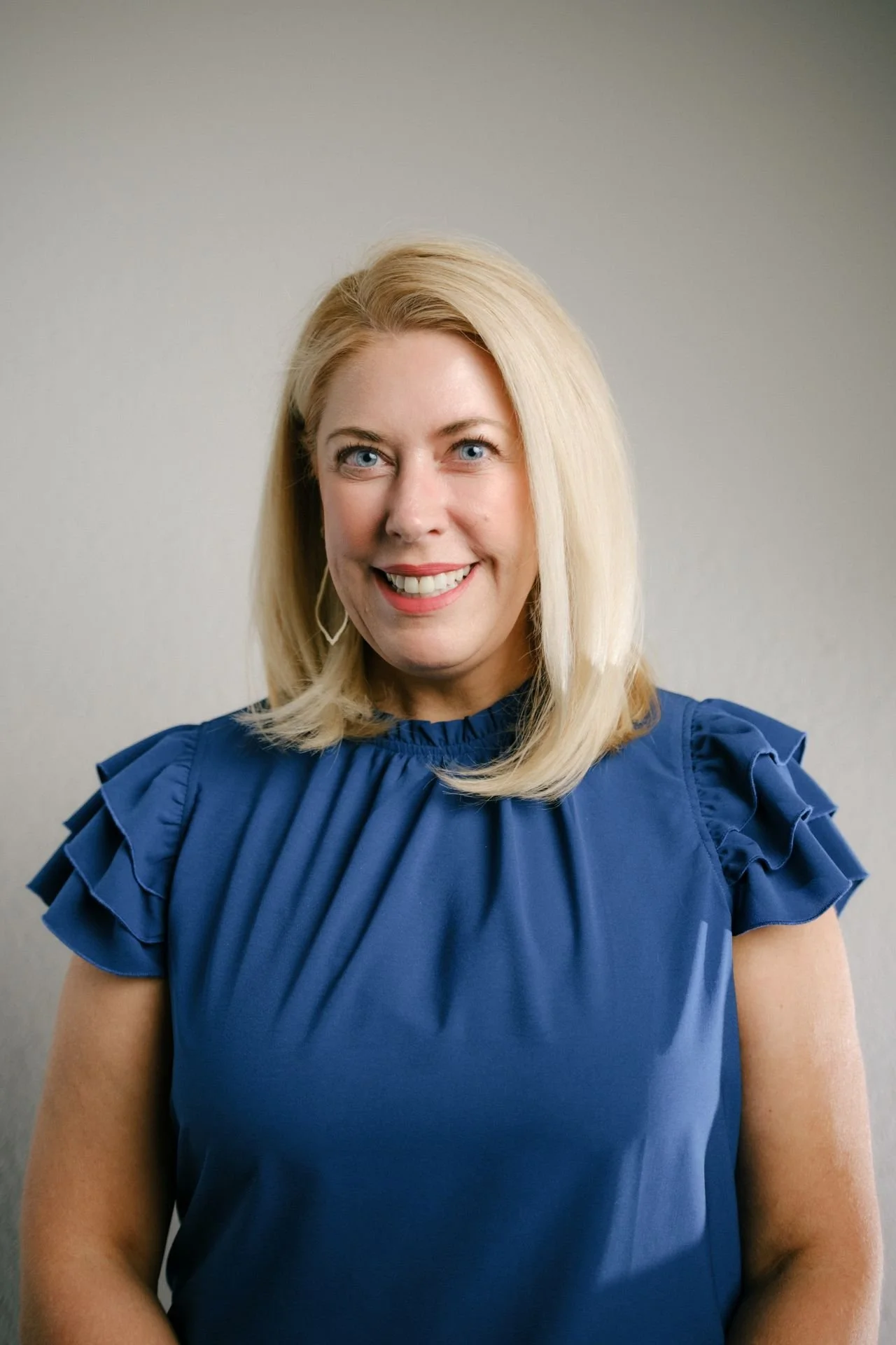 Woman with shoulder length straight blonde hair wearing a blue top with ruffly sleeves
