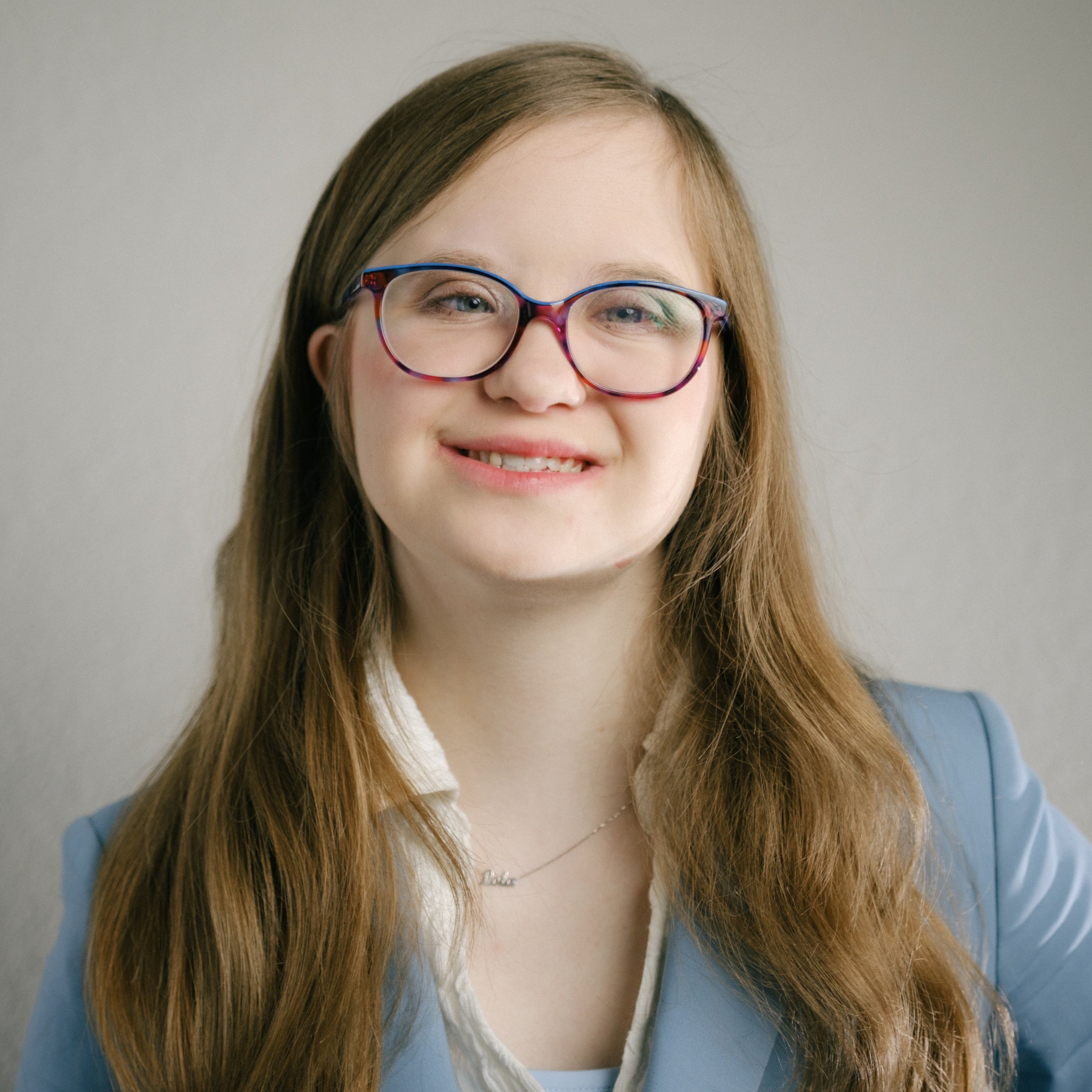 Woman with long blonde avy hair wearing a gray suit and glasses