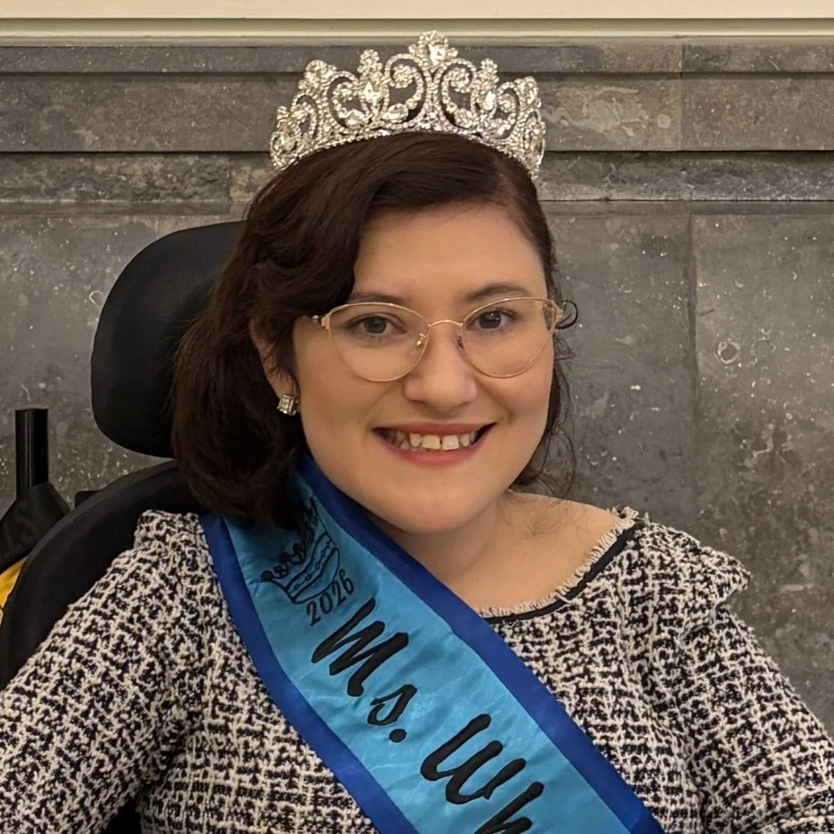 White woman with dark wavy hair pulled to once side. She is wearing glasses and a tiara and a blue sash over a black and white houndstooth top