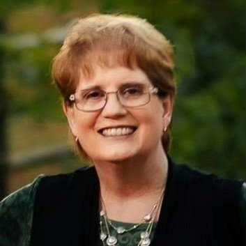 White woman with red hair wearing glasses and a dark green top
