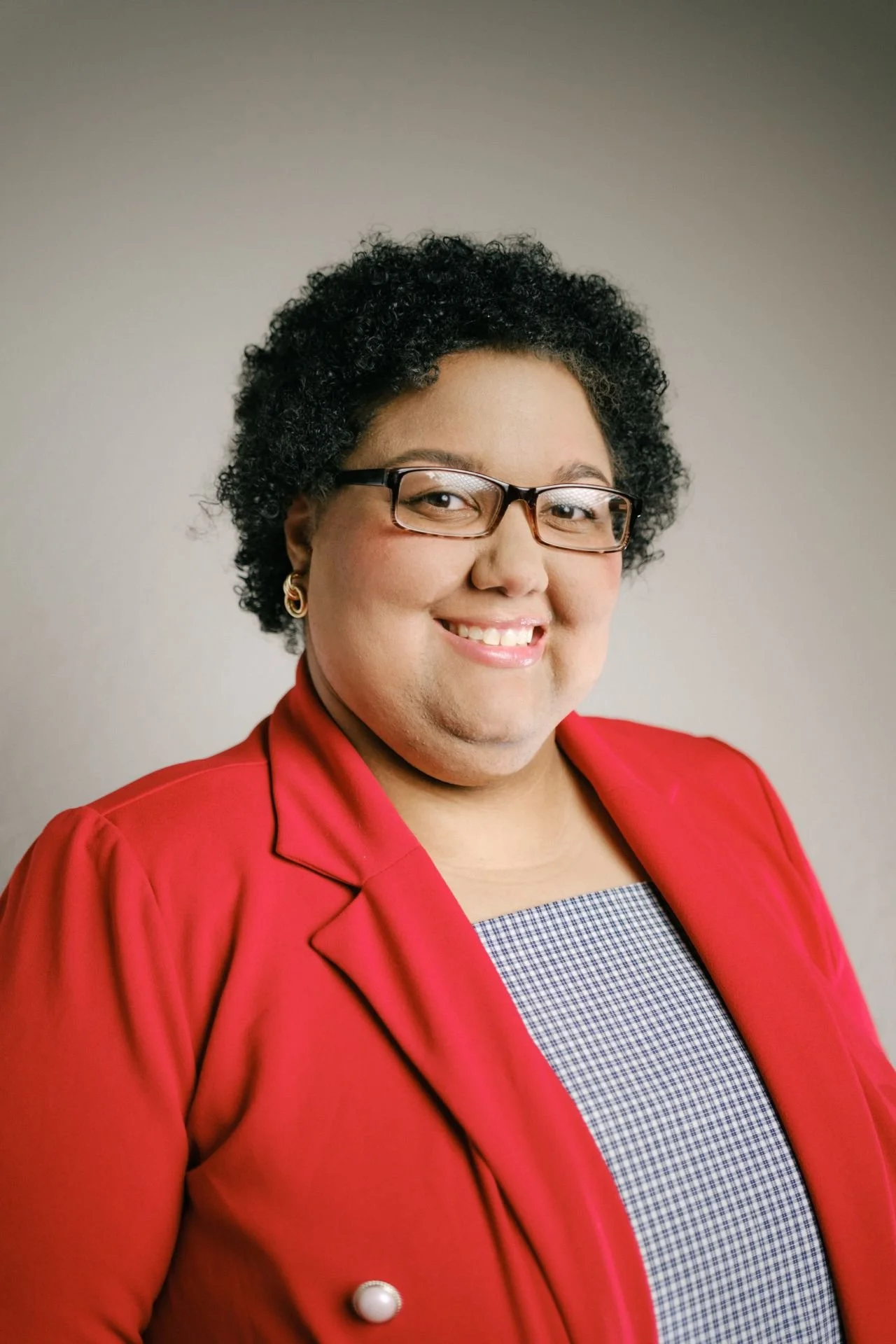 Black woman with curly black hair wearing glasses and a red suit jacket over a gray top
