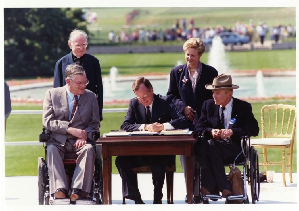 Four people are standing/sitting around a table with green grass and a fountain in the background. A man is sitting behind the table with some paperwork in front of him and is signing one of them with a pen in his hand.
