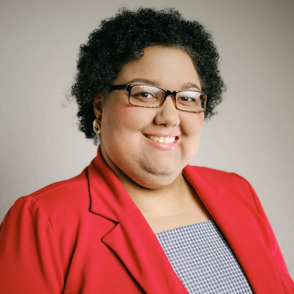 Black woman with curly black hair wearing glasses and a red suit jacket over a gray top