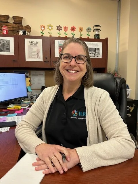 Woman with short light brown hair wearing a black shirt that says ILRC and a white sweater