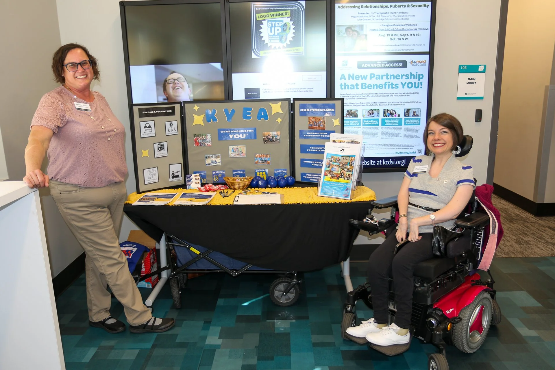 A woman with short hair wearinga gray and blue striped top and black pants using a wheelchair sitting to the right of a table with a KYEA display
