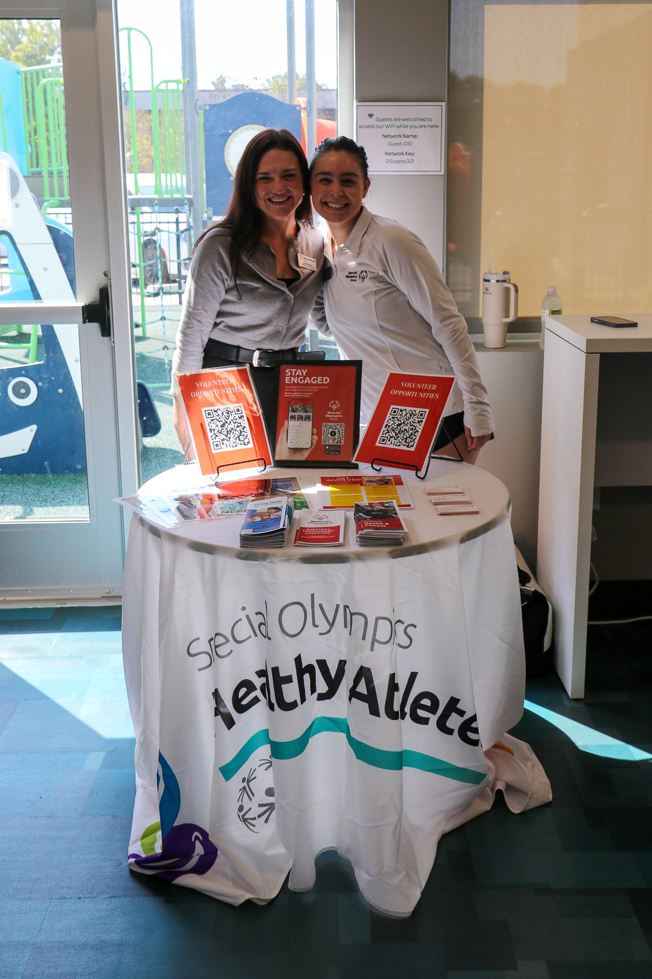 Two women with dark hair wearing white tops behind a table with a white banner Special Olympics Health Athletes