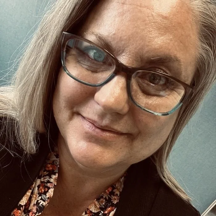 Woman with blonde shoulder length hair wearing a printed top with a black sweater and glasses