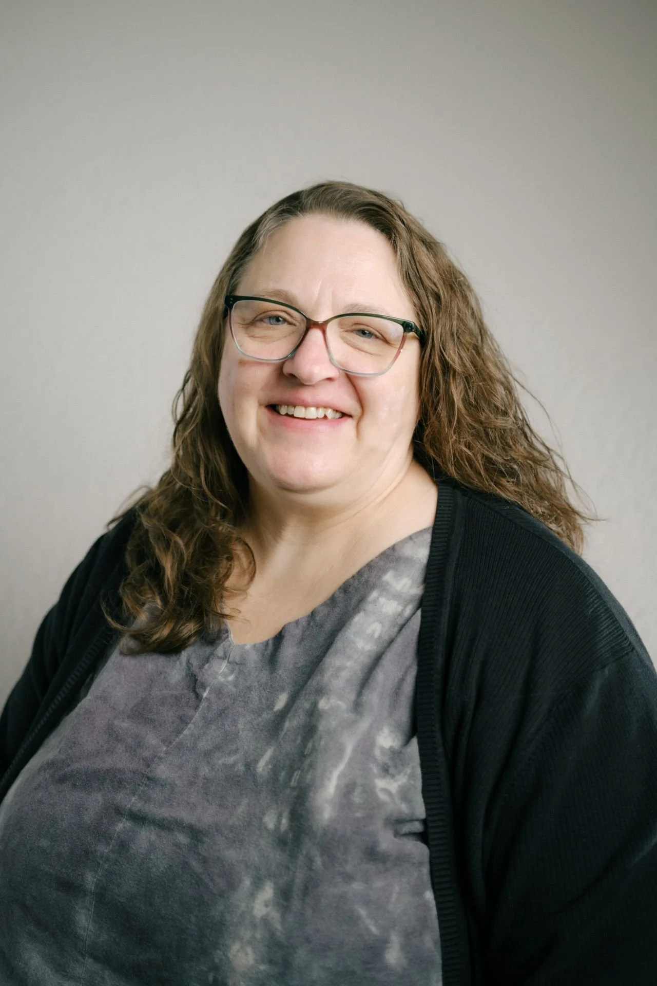 Woman with light brown long dark curly hair wearing glasses and a gray top with a black cardigan