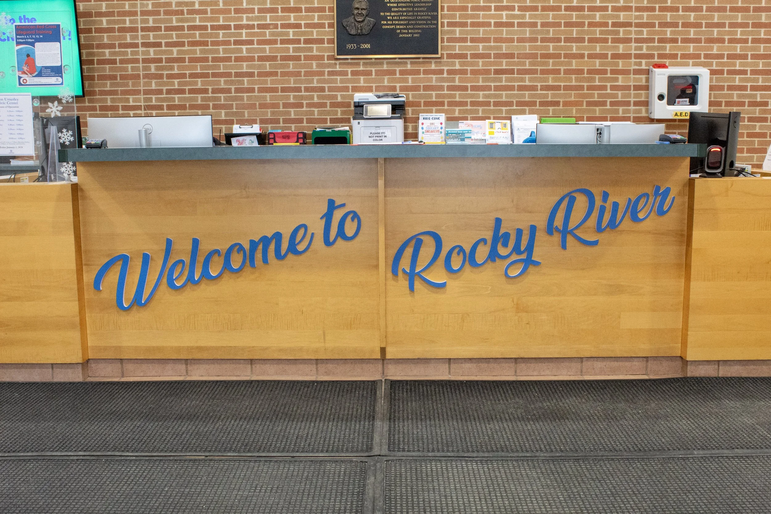 A wooden reception desk with the sign 'Welcome to Rocky River' in blue cursive letters. Behind the desk is a brick wall with a memorial plaque and various office supplies, including a computer, a printer, and a AED box.