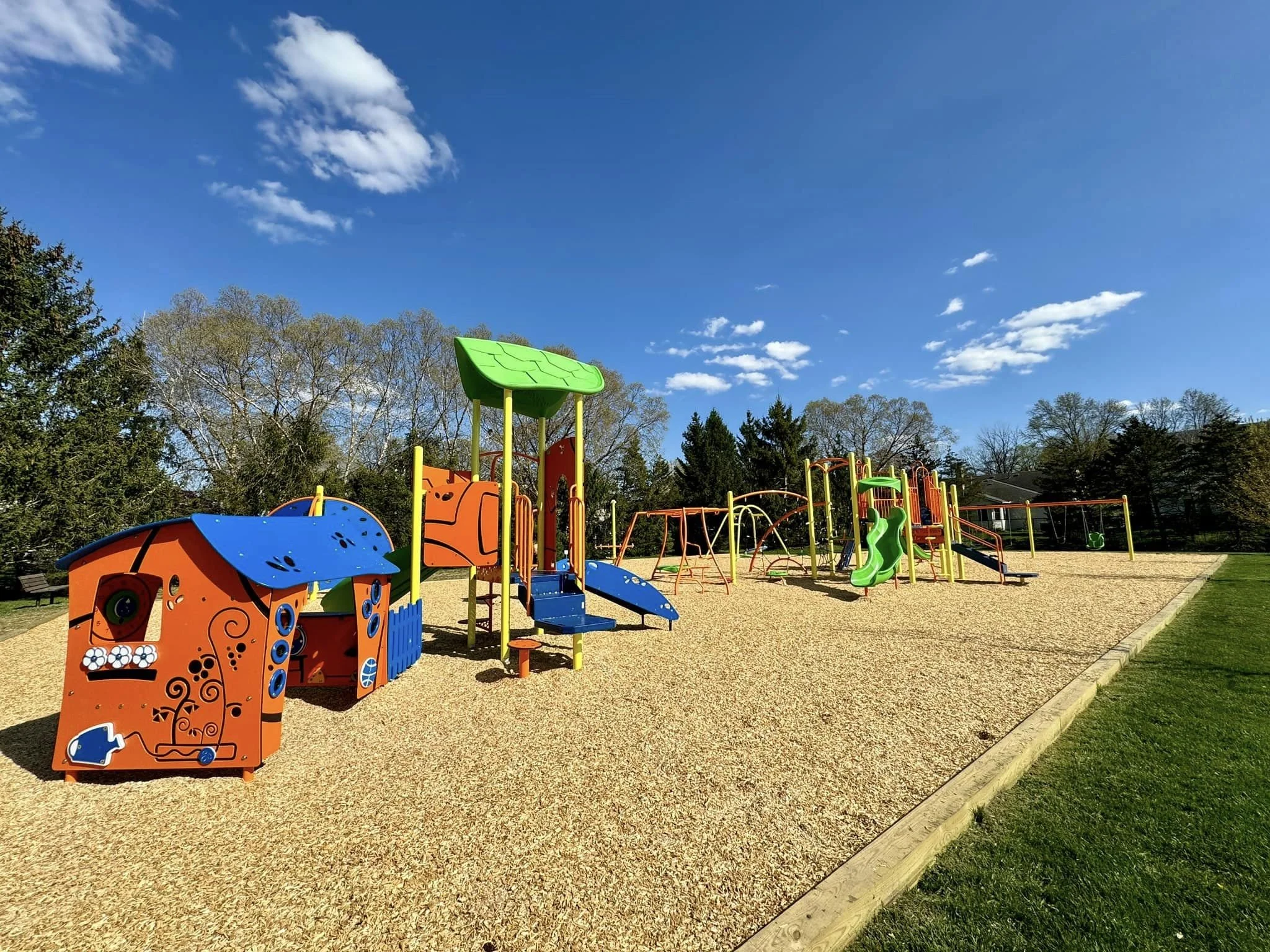 Colorful playground with slides, swings, and climbing structures in a park, under a blue sky with some clouds and trees in the background.