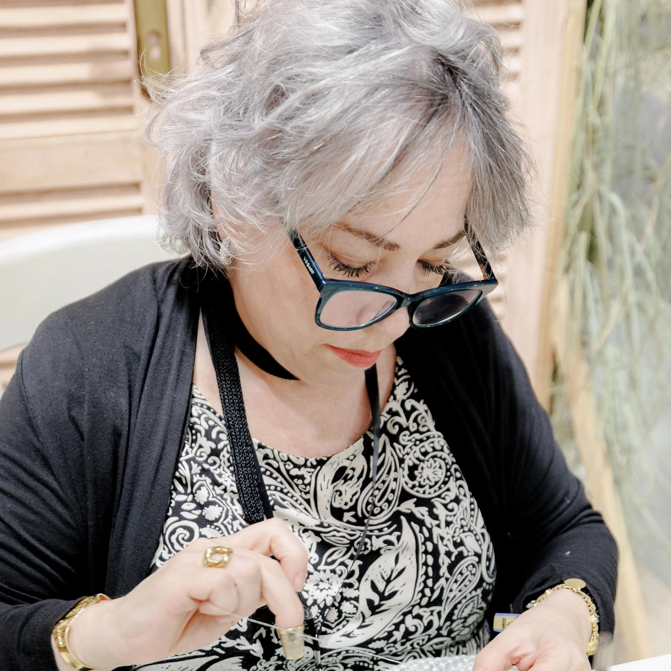 Mujer mayor con cabello canoso y gafas leyendo o escribiendo mientras sostiene un bolígrafo, con un vestido estampado en blanco y negro.