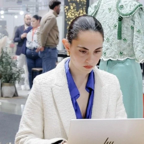 Mujer trabajando en una computadora portátil en un entorno elegante con un maniquí y personas al fondo.