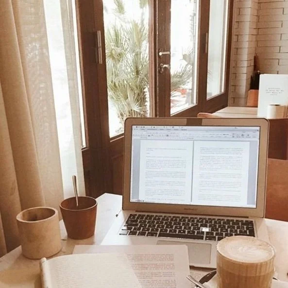 Mesa de café con computadora portátil, documentos, taza de café y planta al fondo.