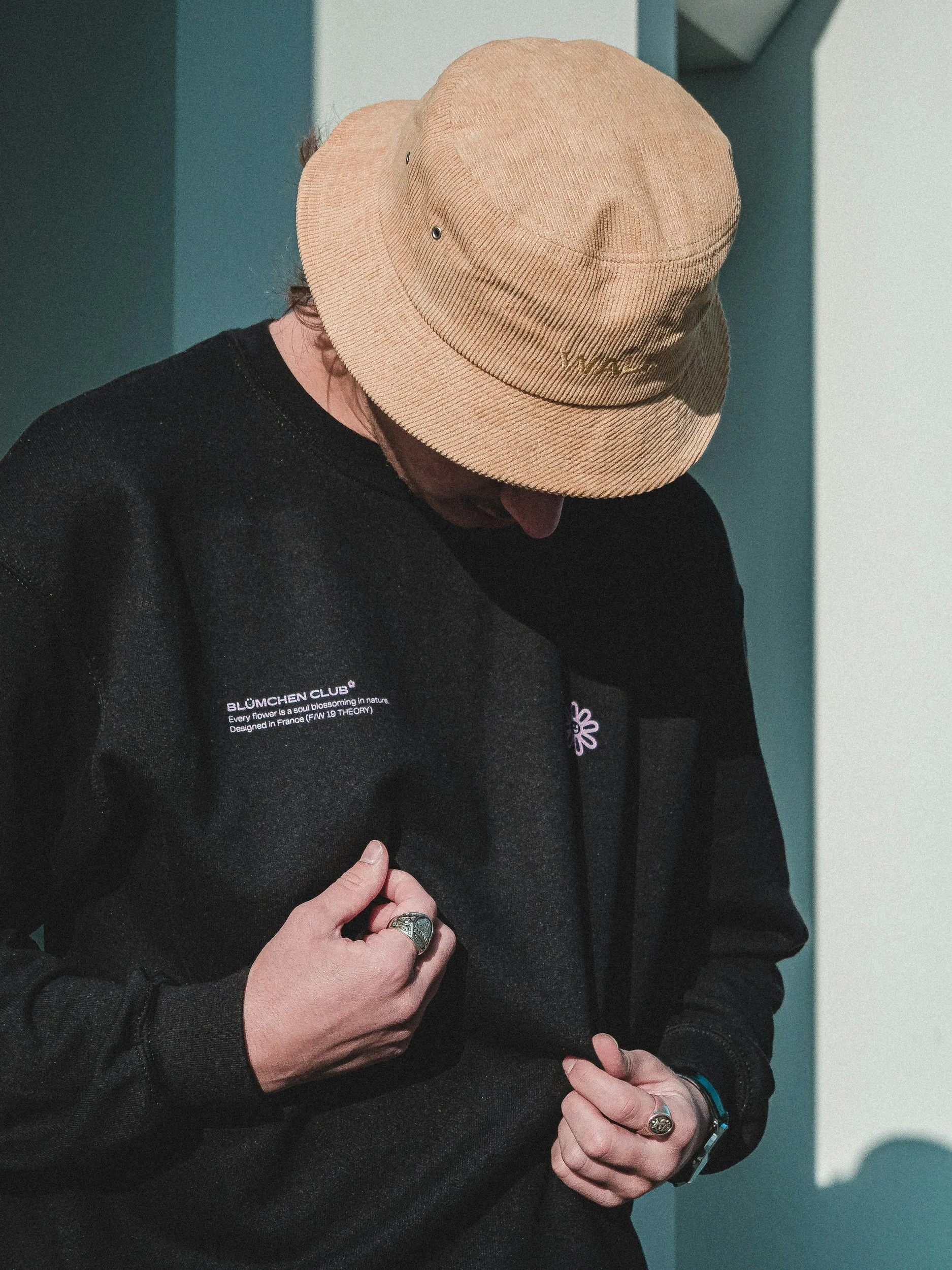 Person wearing a beige bucket hat and black sweatshirt, looking down and adjusting the sweatshirt with their hands.