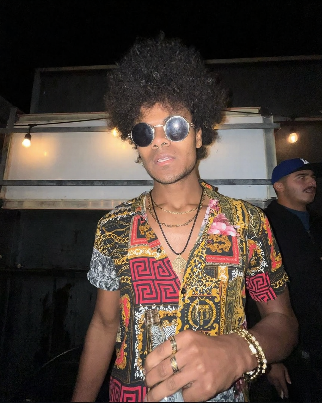 Person with curly hair wearing a black hat with a yellow pixelated dollar sign on it, covering part of their face with their hand.